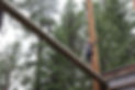 Image taken from below of a large horizontal pole in the air. A camper wearing a harness has her hands extended and is carefully walking across the pole.